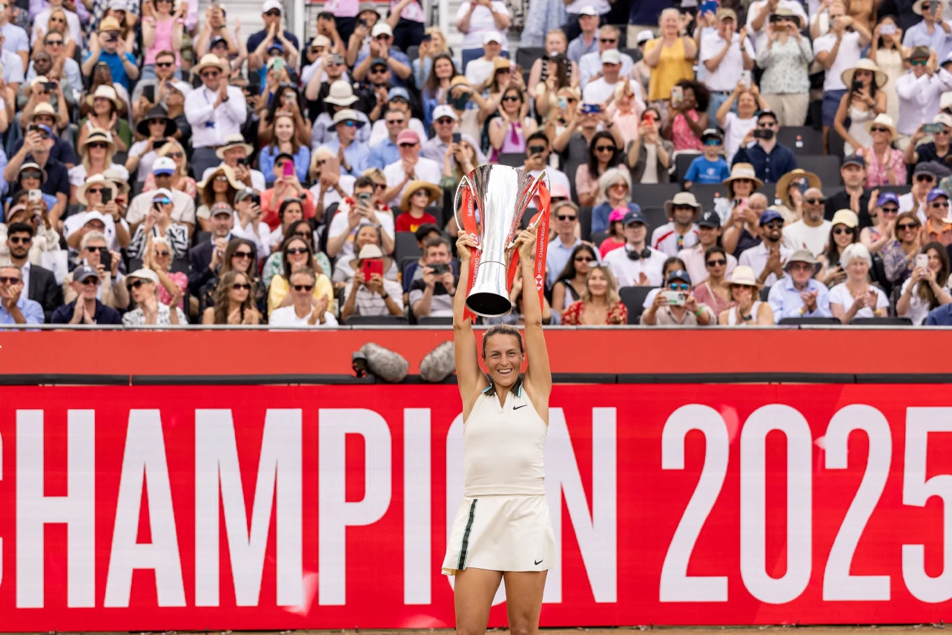 Zawodniczka w stroju tenisowym unosi nad głową trofeum, stojąc na tle dużego napisu 'CHAMPION 2025', za nią tłum wiwatujących kibiców.