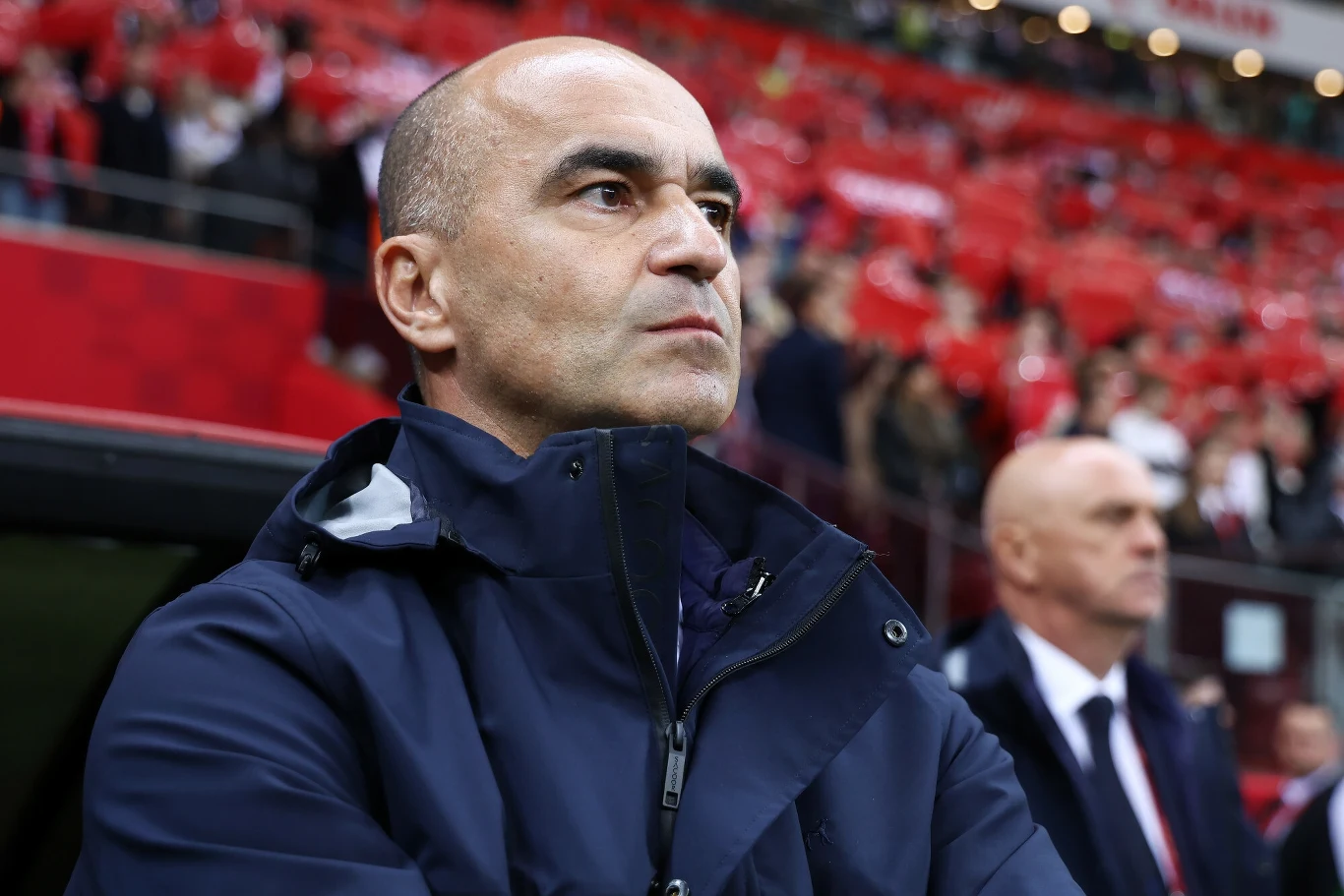 Roberto Martinez Mężczyzna w kurtce sportowej patrzący skupionym wzrokiem, w tle widać trybuny stadionu i drugą osobę ubraną elegancko.