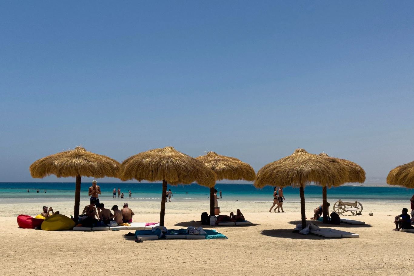 Marsa Alam to kurort w Egipcie, nad Morzem Czerwonym Plaża z jasnym piaskiem, na której wypoczywają ludzie pod dużymi słomianymi parasolami, w tle błękitne morze i niebo bez chmur.