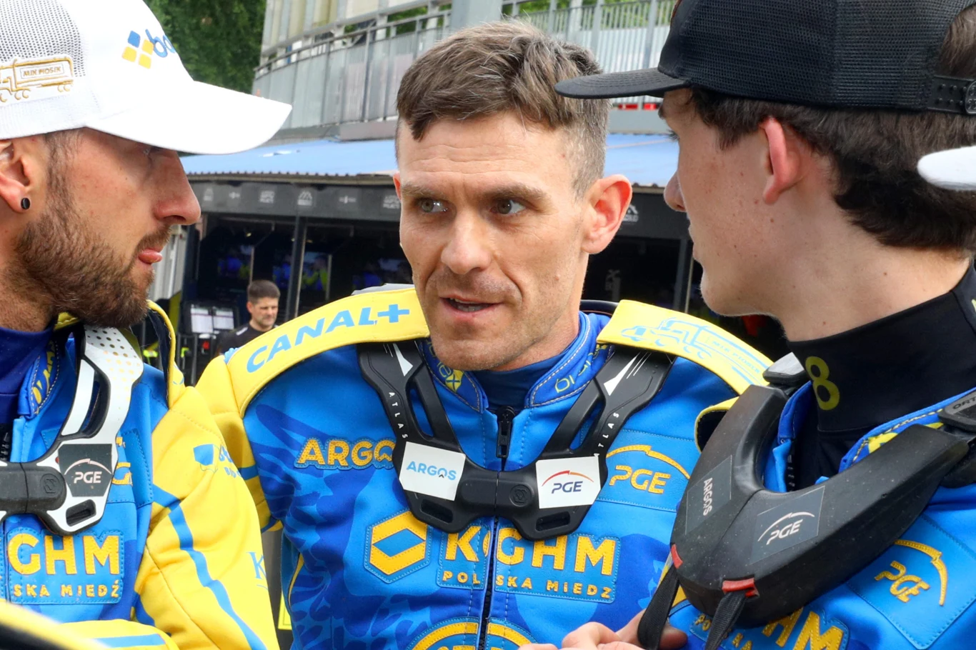 Martin Vaculik w rozmowie z Andersem Thomsenem. Trzech żużlowców w strojach sportowych z logo sponsorów, stojących blisko siebie i rozmawiających, w tle widoczne elementy toru oraz osoby z obsługi.