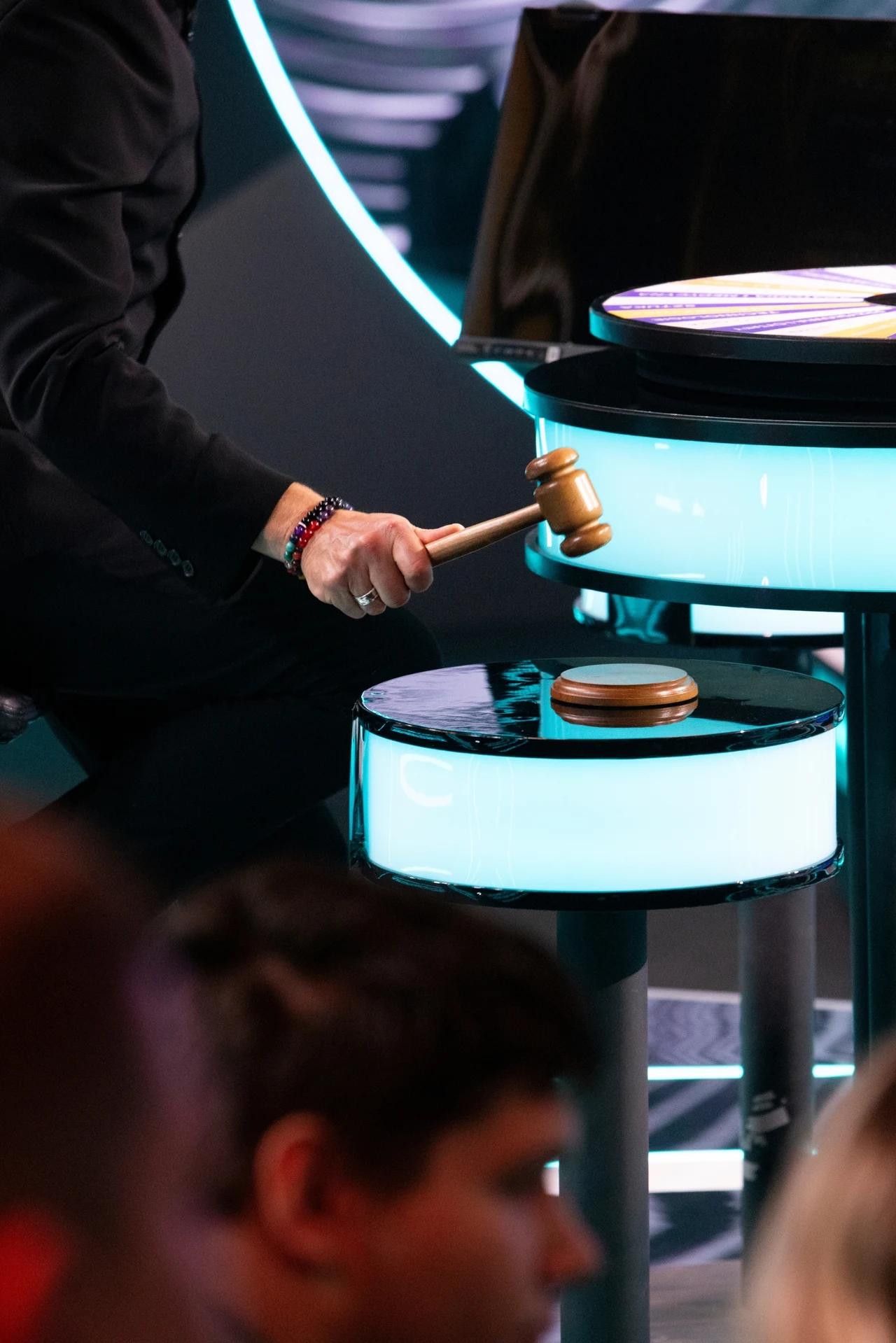 Osoba w garniturze uderza drewnianym młotkiem o okrągłą podstawkę na jasno oświetlonym stole, co sugeruje scenę związaną z aukcją lub podejmowaniem decyzji.