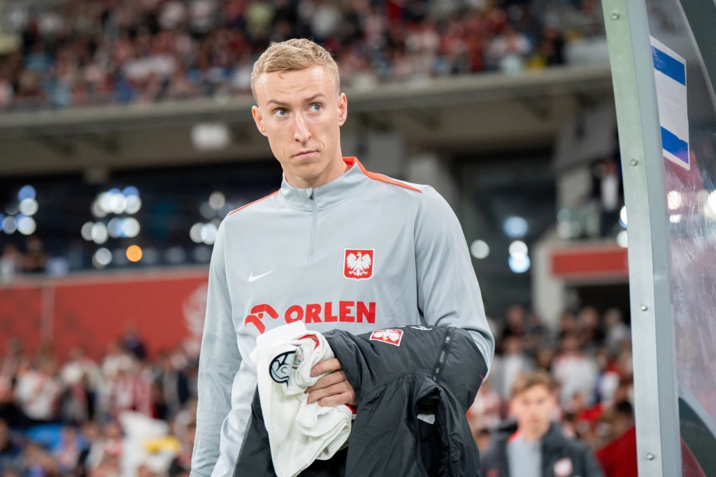 Adam Buksa Mężczyzna w szarej sportowej bluzie z godłem Polski oraz napisem Orlen stoi na stadionie z kurtką przewieszoną przez rękę, w tle rozmyta publiczność.