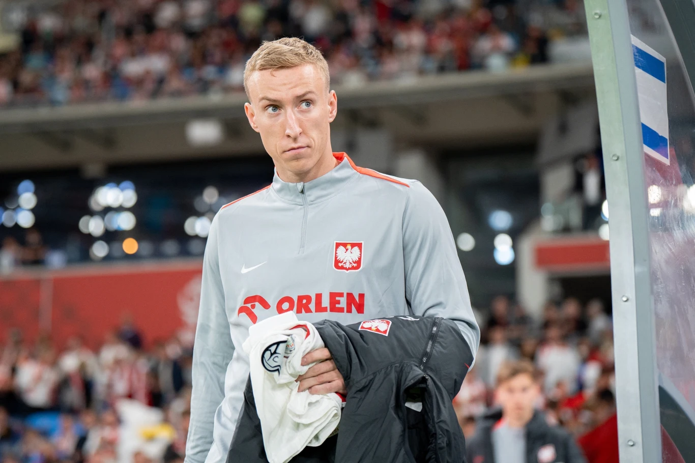 Mężczyzna w szarej sportowej bluzie z godłem Polski oraz napisem Orlen stoi na stadionie z kurtką przewieszoną przez rękę, w tle rozmyta publiczność.