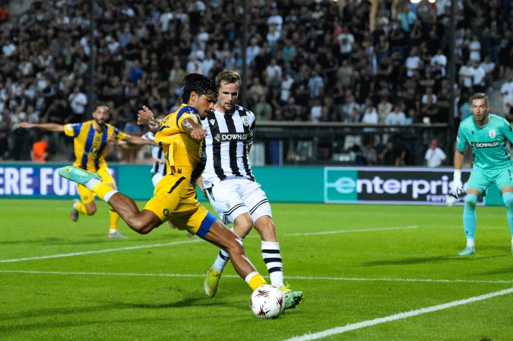 Zawodnik w żółtym stroju wykonuje dynamiczne kopnięcie piłki przy próbie wyprzedzenia defensora drużyny w czarno-białych pasach na stadionie pełnym kibiców, obok bramkarz uważnie obserwuje sytuację.