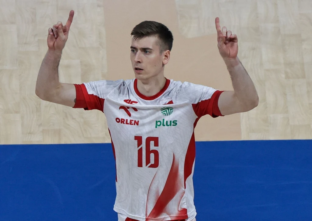 Kamil Semeniuk Siatkarz w białoczerwonym stroju z numerem 16 unosi obie ręce do góry, stojąc na boisku sportowym. Na koszulce widoczne są logotypy sponsorów Orlen oraz Plus.