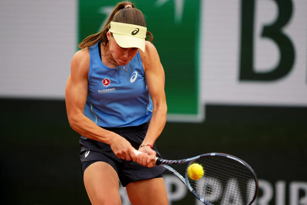 Tenisistka podczas intensywnego uderzenia piłki rakietą, w sportowym stroju, skupiona na grze, w tle widoczna część kortu i elementy oznakowania turnieju.