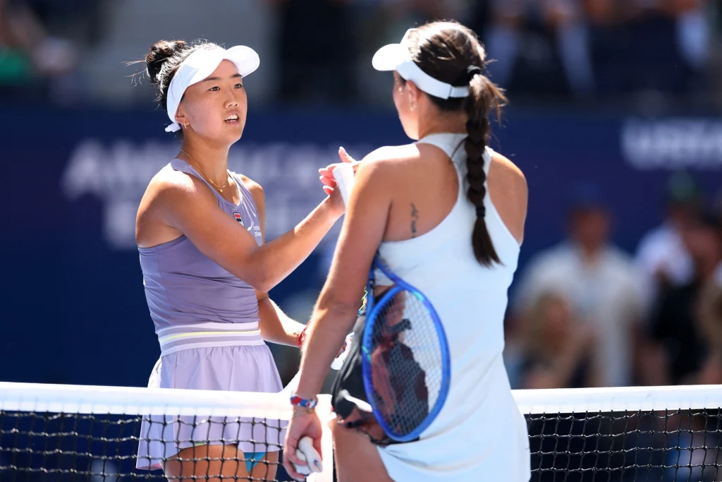 Ann Li Dwie tenisistki stoją naprzeciwko siebie przy siatce kortu tenisowego, wymieniając uścisk dłoni po meczu, obie ubrane w sportowe stroje, w tle rozmyta widownia.