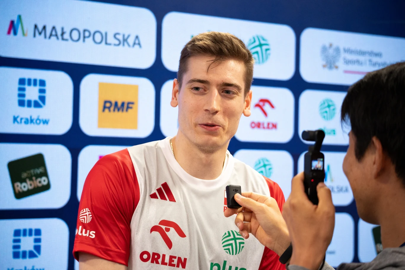 Zawodnik w stroju sportowym udzielający wywiadu dziennikarzowi, w tle ścianka z logotypami sponsorów oraz instytucji związanych z wydarzeniem.