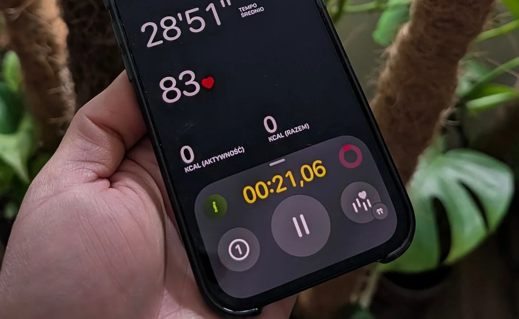 Dłoń trzymająca telefon z aplikacją monitorującą aktywność fizyczną, widoczny stoper, tętno 83, czas trwania treningu ponad 28 minut oraz licznik spalonych kalorii ustawiony na zero, w tle liście roślin.