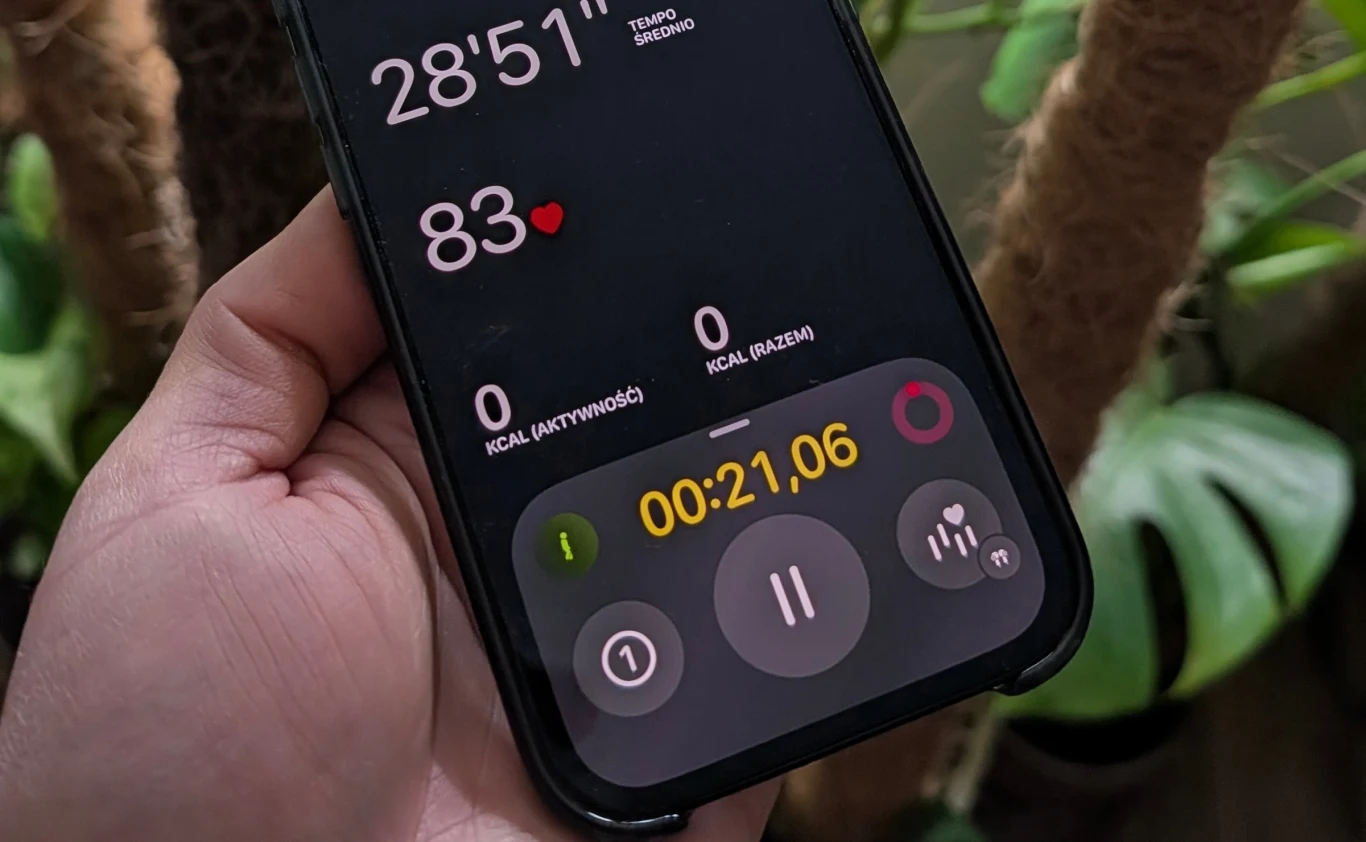Dłoń trzymająca telefon z aplikacją monitorującą aktywność fizyczną, widoczny stoper, tętno 83, czas trwania treningu ponad 28 minut oraz licznik spalonych kalorii ustawiony na zero, w tle liście roślin.