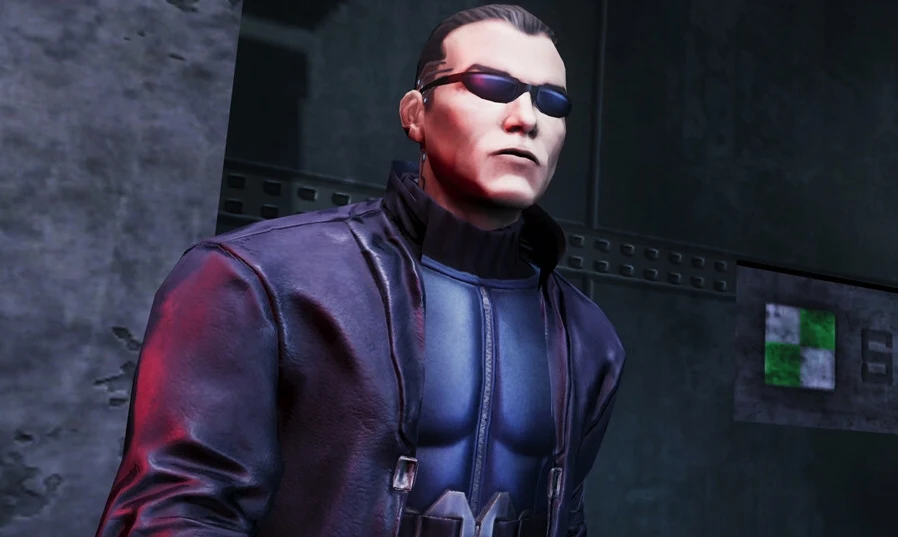 Deus Ex Remastered Mężczyzna o krótkich ciemnych włosach, w czarnych okularach przeciwsłonecznych, ubrany w ciemną skórzaną kurtkę i niebieski kombinezon, stoi na tle industrialnej ściany.