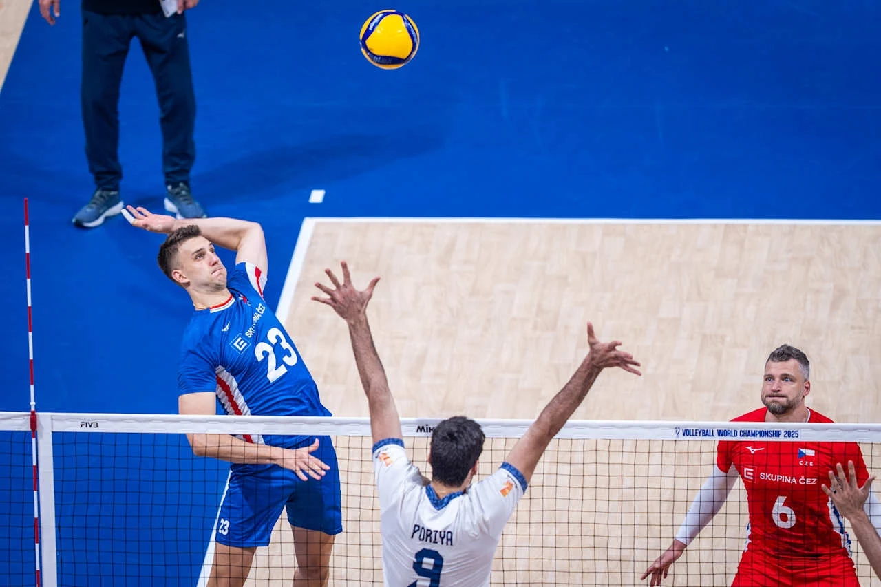 Zawodnik w niebieskim stroju unosi się w powietrzu do ataku siatkarskiego, naprzeciw niego wyciąga ręce blokujący w białym stroju, w tle drugi zawodnik w czerwonym stroju obserwuje akcję; piłka siatkarska znajduje się nad siatką.