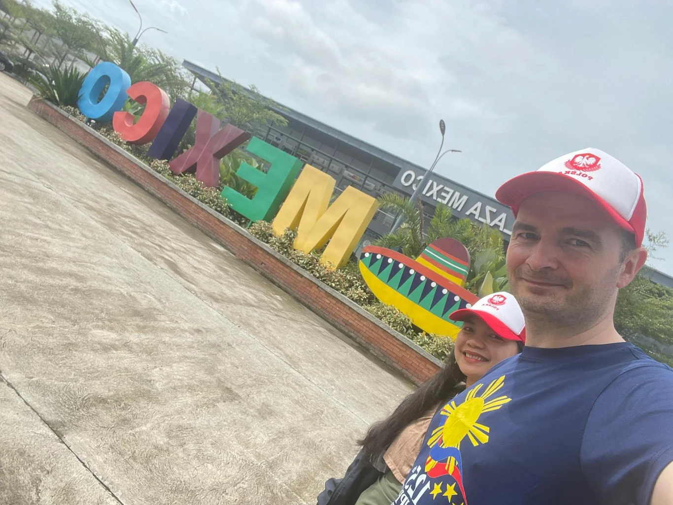 Malou i Andrzej w Mexico, w prowincji Pampanga, skąd pochodzi Filipinka Mężczyzna i kobieta w czapkach z daszkiem pozują do selfie na tle kolorowego napisu "MEXICO" oraz dekoracyjnego sombrero, w otoczeniu roślin i nowoczesnego budynku.