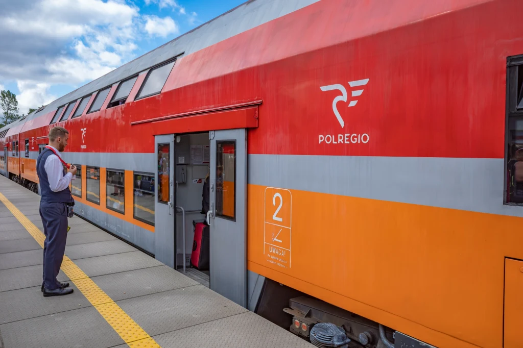 Dwupiętrowy pociąg pasażerski Polregio stojący przy peronie z otwartymi drzwiami, obok którego stoi konduktor w mundurze, w tle błękitne niebo z chmurami.