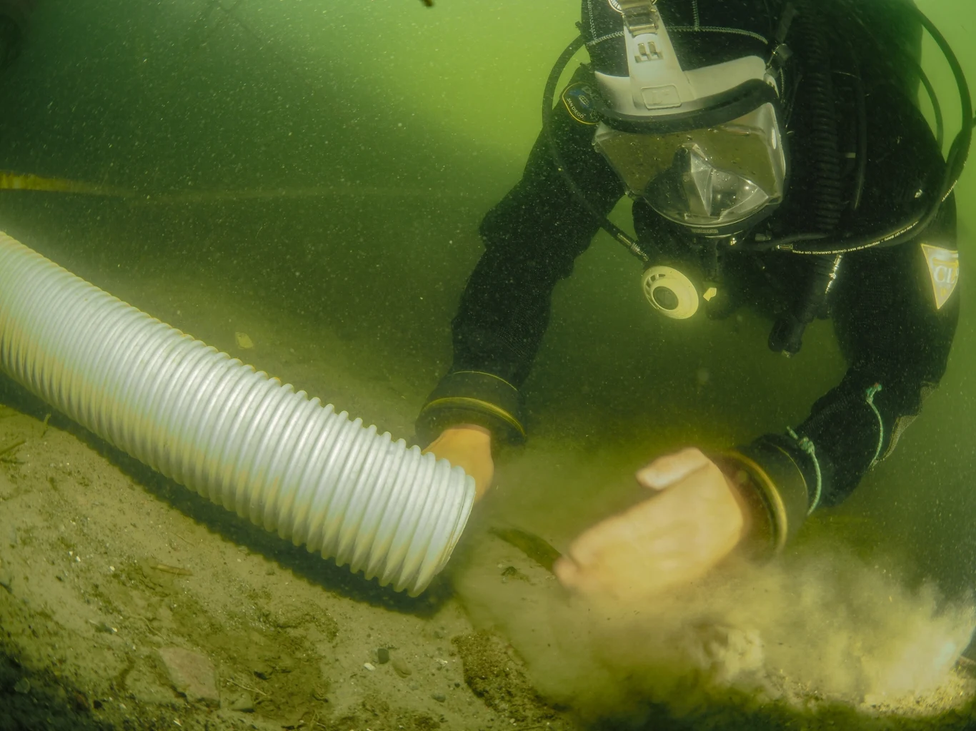 Archeolodzy podwodni wykorzystują wiele metod do badań. Zdjęcie autorstwa Marcina Trzcińskiego z badań portu w Pucku. Dzięki uprzejmości dr. Mateusza Popka. Nurek w specjalistycznym sprzęcie pracuje pod wodą, przeszukując dno i używając elastycznego węża do odsysania piasku lub osadów.