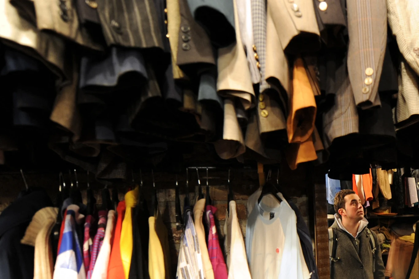 Wnętrze sklepu z ubraniami, na pierwszym planie liczne marynarki i płaszcze zawieszone na wieszakach pod sufitem, w tle mężczyzna przyglądający się asortymentowi.