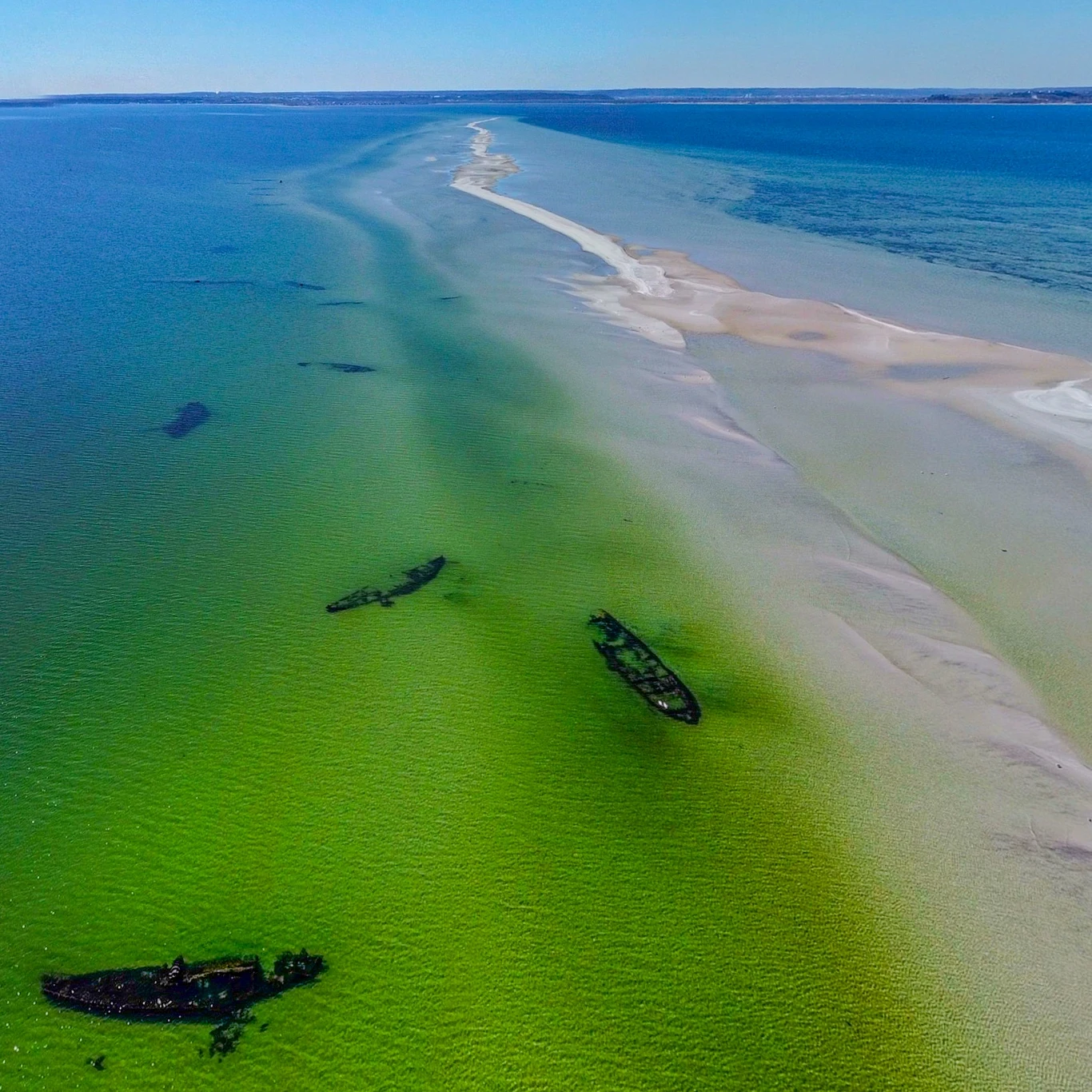 Wrakowisko przy Rewie Mew. Na mieliźnie znajduje się ich kilkadziesiąt. Najlepiej widać to z drona. Wraki statków zanurzone w płytkiej, turkusowej wodzie przy piaszczystej mieliźnie, z rozciągającym się morskim horyzontem pod błękitnym niebem.