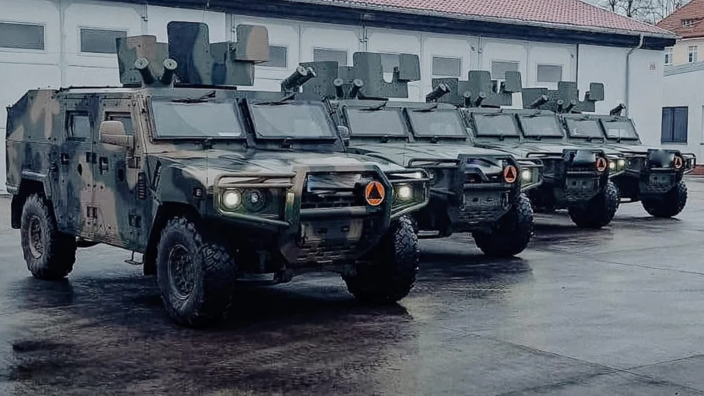 Cztery wojskowe pojazdy opancerzone ustawione w szeregu na placu, każdy z charakterystycznymi emblematami ochrony cywilnej na masce i specjalistycznym wyposażeniem na dachu, w tle budynek z białą elewacją.