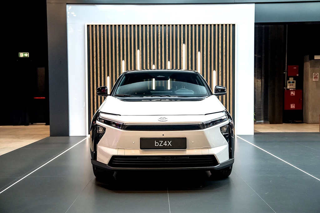 Nowoczesny, biały samochód elektryczny Toyota bZ4X ustawiony centralnie w przestronnym, eleganckim wnętrzu z geometrycznym tłem w postaci drewnianych paneli i dużego, jasnego obramowania.