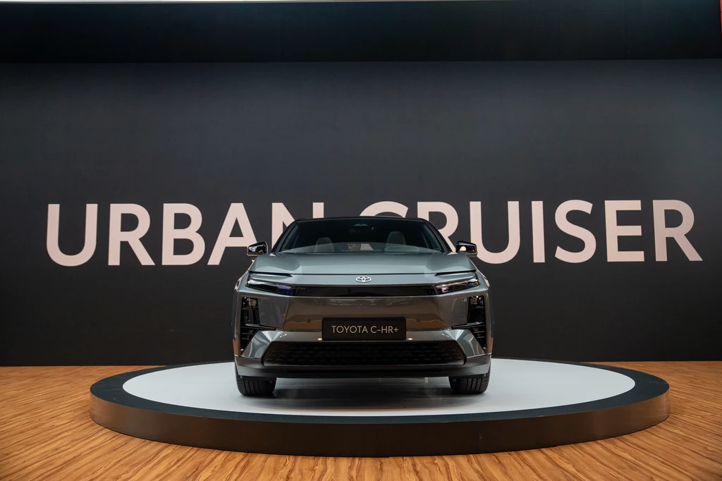 Nowoczesny samochód Toyota C-HR ustawiony na podświetlonej platformie, na tle czarnej ściany z dużym białym napisem Urban Cruiser, prezentowany w eleganckiej przestrzeni wystawowej.