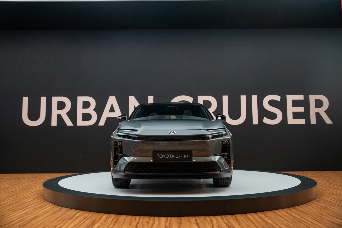 Nowoczesny samochód Toyota C-HR ustawiony na podświetlonej platformie, na tle czarnej ściany z dużym białym napisem Urban Cruiser, prezentowany w eleganckiej przestrzeni wystawowej.