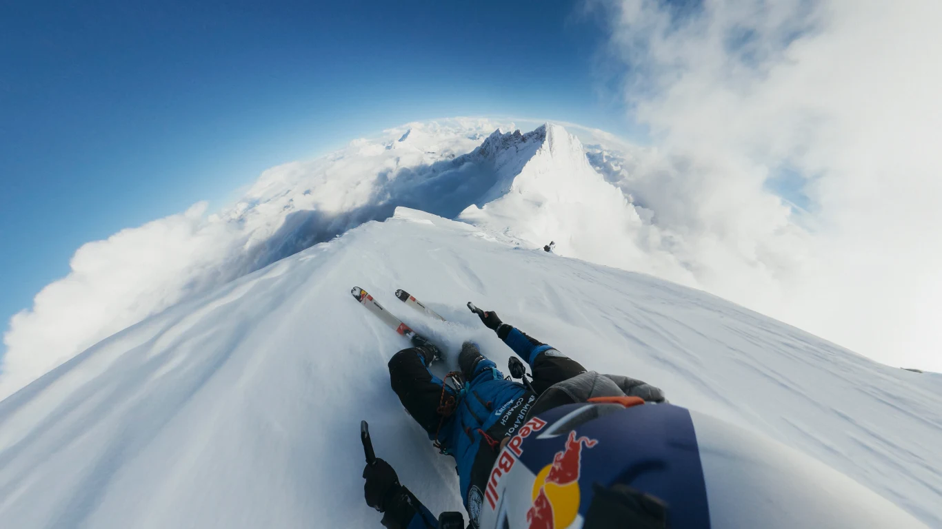 Narciarz w specjalistycznym kombinezonie i kasku z widocznym logo Red Bull zjeżdża stromym zboczem ośnieżonej góry, wokół roztacza się imponujący krajobraz gór i chmur pod błękitnym niebem.