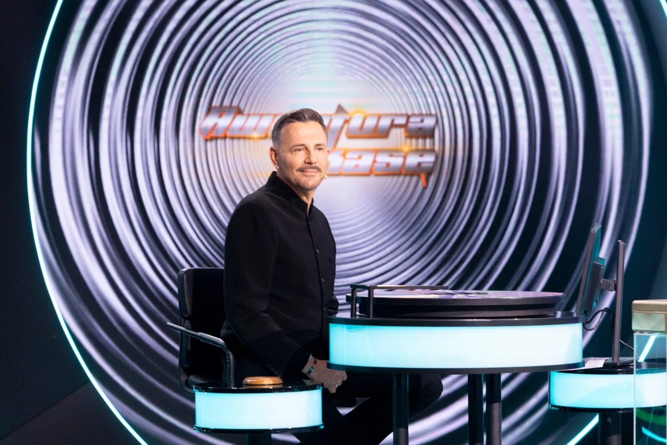 Awantura o kasę. Trening wiedzy o 17:30 w niedzielę w Polsacie Mężczyzna siedzący przy nowoczesnym, podświetlanym biurku w studiu telewizyjnym, w tle okrągła grafika z napisem i tunelem optycznym, profesjonalna sceneria programu lub konkursu telewizyjnego.