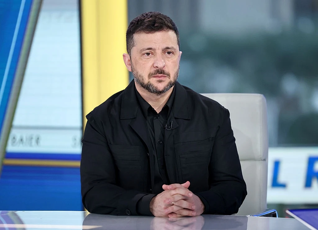 Wołodymyr Zełenski Mężczyzna w ciemnym ubraniu siedzący przy biurku w studiu telewizyjnym, z poważnym wyrazem twarzy, dłonie splecione na stole, w tle rozmyty ekran.