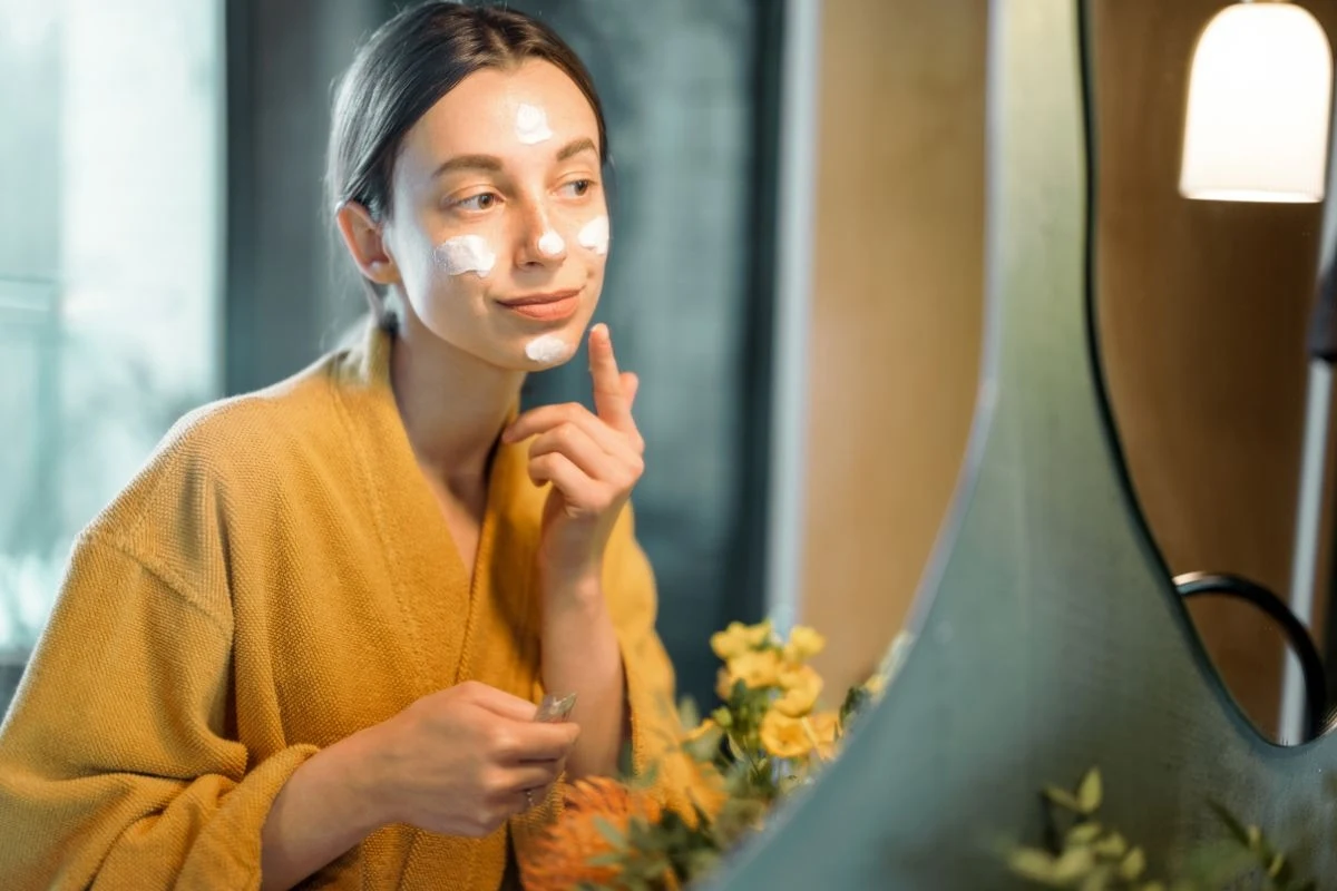 Kobieta w żółtym szlafroku nakłada krem na twarz, stojąc przed lustrem w jasnym, przytulnym pomieszczeniu.