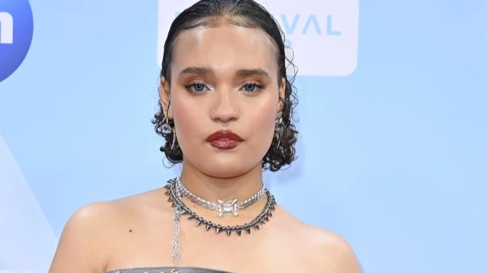 Sara James Młoda kobieta z krótkimi, ciemnymi falowanymi włosami w eleganckim makijażu, ubrana w srebrną kreację z ozdobną biżuterią na szyi, na niebieskim tle z niewyraźnymi napisami w tle.