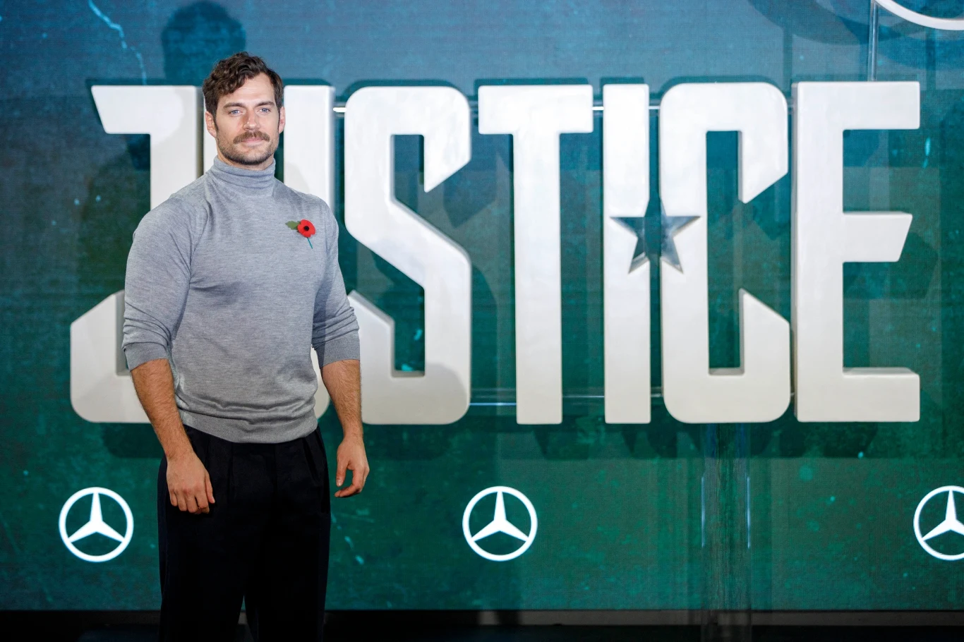 Henry Cavill z wąsem Mężczyzna w szarej golfie z przypiętym czerwonym makiem po lewej stronie stoi na tle dużego napisu JUSTICE i symboli Mercedesa.