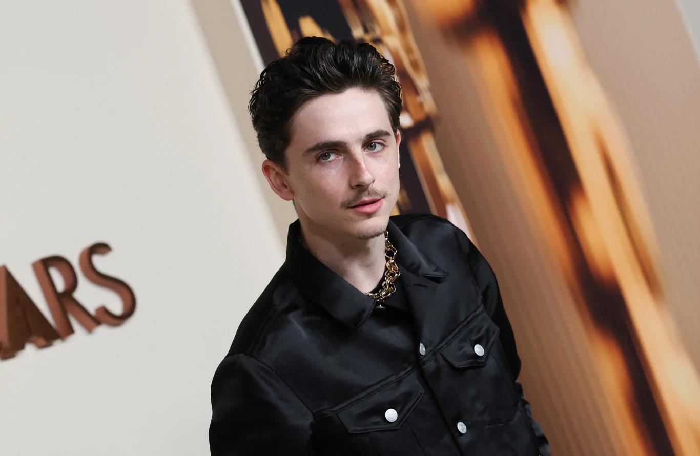 Timothee Chalamet i jego wąsik Mężczyzna w czarnej koszuli i złotym naszyjniku pozuje na tle jasnej ścianki z elementami dekoracyjnymi i napisem, z wyraźnym światłem skierowanym na twarz.