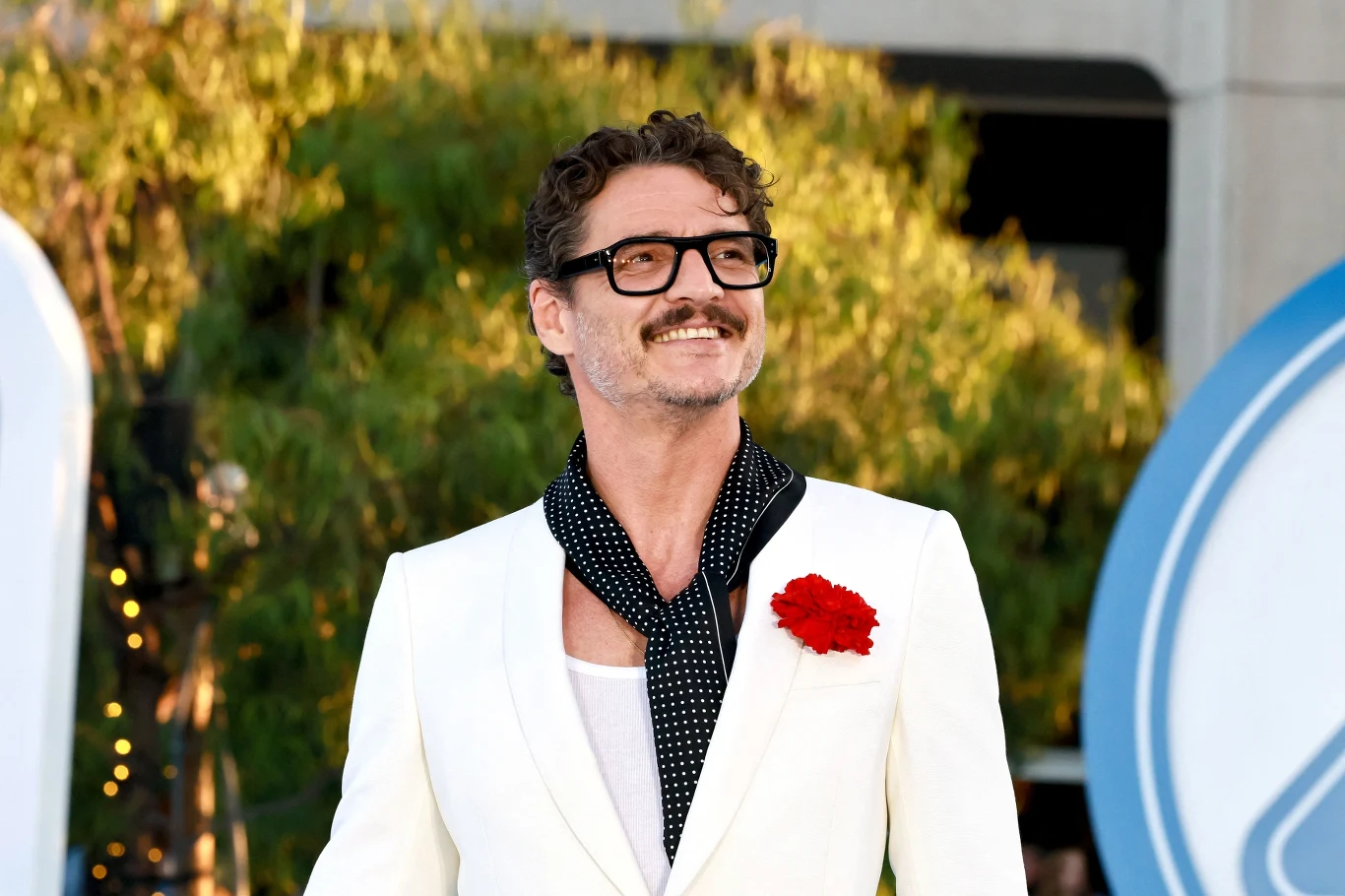 Pedro Pascal Mężczyzna w białym garniturze z czerwoną ozdobą przypiętą do klapy, uśmiechnięty, z ciemnymi okularami i fantazyjnym szalikiem, stoi na tle zieleni oraz nowoczesnego budynku.