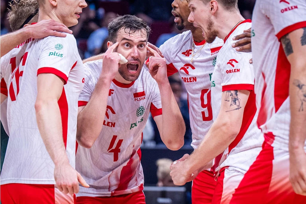 Grupa siatkarzy w biało-czerwonych strojach sportowych z logo sponsorów, skoncentrowanych na rozmowie podczas meczu, z jednym z zawodników energicznie gestykulującym i skupiającym uwagę kolegów.