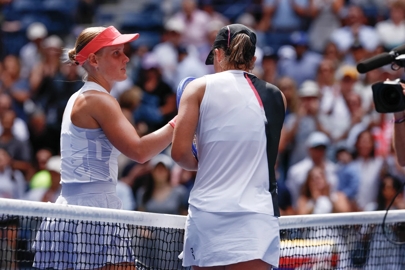 Dwie tenisistki stoją po przeciwnych stronach siatki podczas zakończenia meczu, ściskając sobie dłonie na korcie, w tle rozmyci kibice na trybunach
