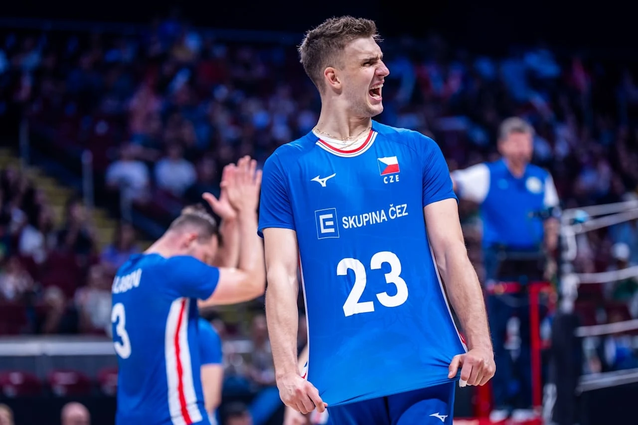 Zawodnik drużyny siatkarskiej w niebieskim stroju z numerem 23 wyraża silne emocje po udanej akcji na boisku, w tle drugi członek zespołu oraz publiczność na trybunach.