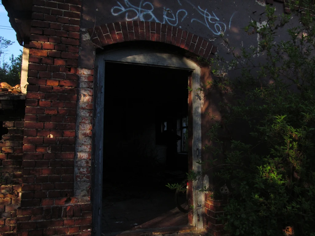 Wejście do opuszczonego, zniszczonego budynku z cegły, otoczonego roślinnością, z widocznym graffiti nad drzwiami i ciemnym wnętrzem w tle.
