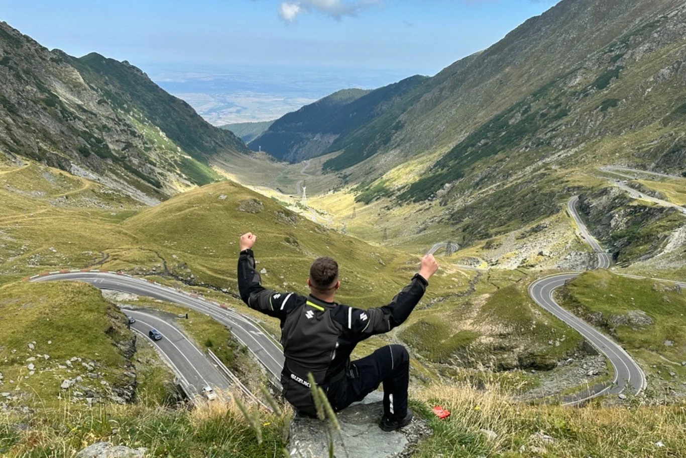 Chcą wprowadzić opłatę za przejazd drogą Transfogaraską w Rumunii. Motocyklista w czarnym kombinezonie siedzi na skale na tle spektakularnej, wijącej się drogi prowadzącej przez górską dolinę, wokół zielone zbocza i otwarta przestrzeń.