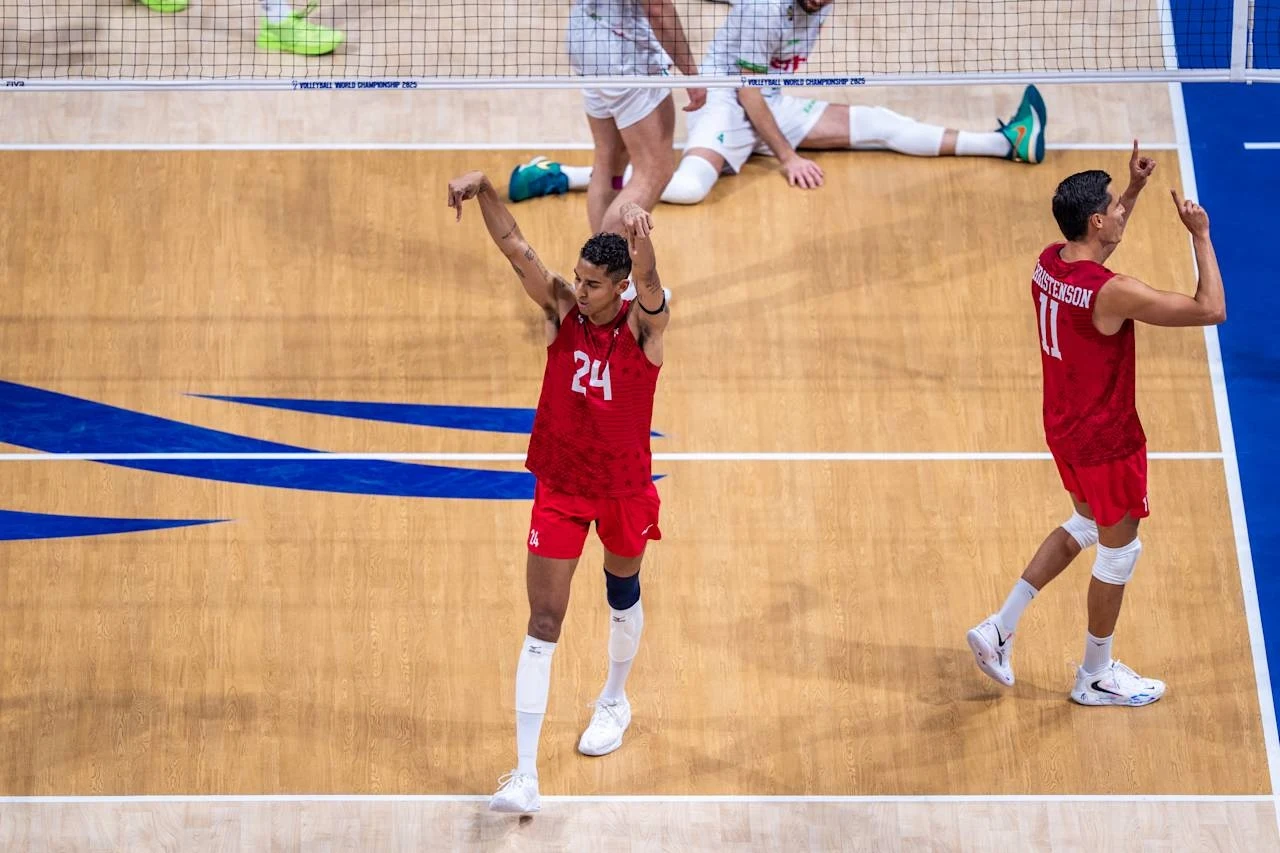 Siatkarze reprezentacji USA Dwóch siatkarzy w czerwonych strojach cieszy się z punktu, unosząc ręce w górę, podczas gdy jeden z przeciwników w białym stroju siedzi na parkiecie, a drugi klęczy tuż za siatką.