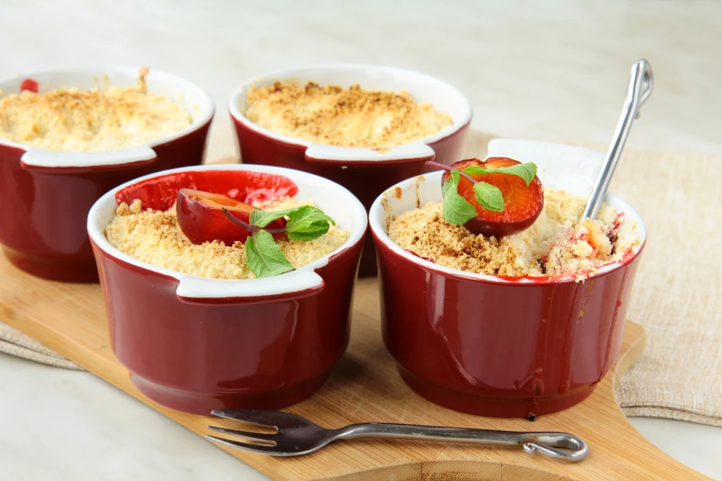 Cztery czerwone kokilki z deserem crumble, każda ozdobiona połówką śliwki i listkami mięty, ustawione na drewnianej desce, obok metalowy widelec.