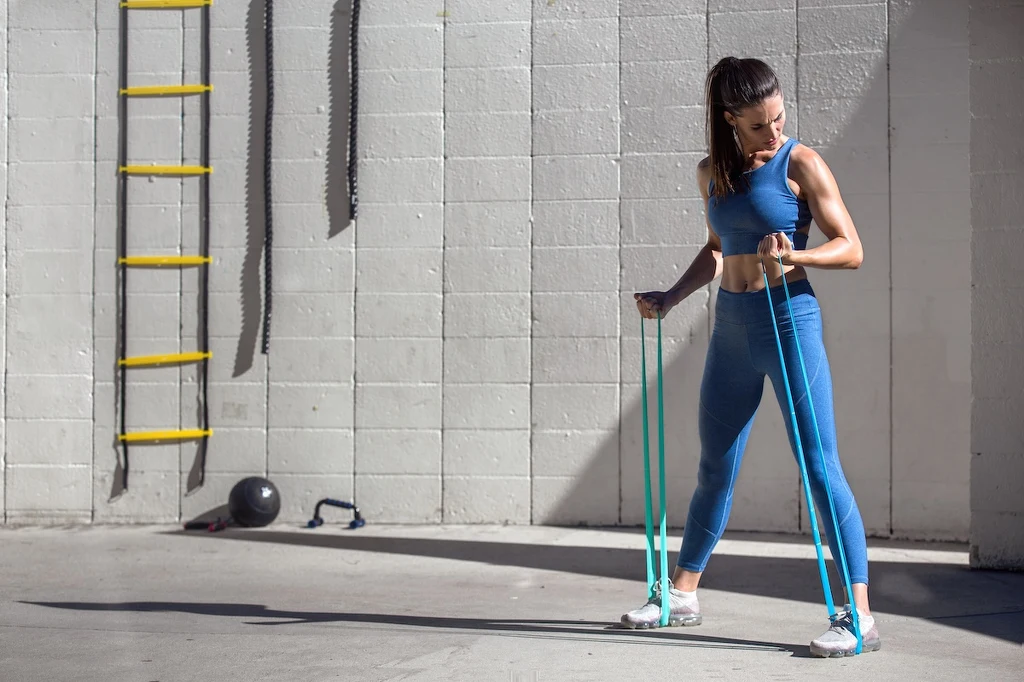 Trening z taśmami 4FIZJO Kobieta w sportowym stroju trenuje z gumą oporową w siłowni, promienie słoneczne wpadają przez okno, a w tle widoczna jest drabinka treningowa i piłka lekarska.