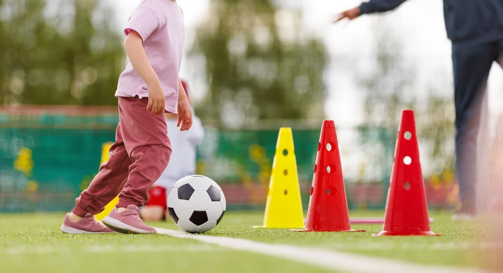 Dziecko w różowym stroju prowadzi piłkę nożną wśród kolorowych pachołków treningowych ustawionych na zielonym boisku.