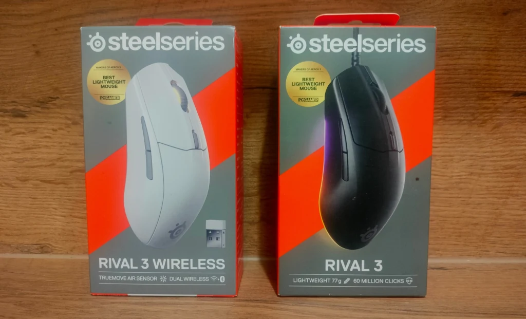 Dwie myszki komputerowe SteelSeries Rival 3 w oryginalnych opakowaniach, jedna w wersji bezprzewodowej w kolorze białym, druga przewodowa w kolorze czarnym, obie prezentowane na tle drewnianym.