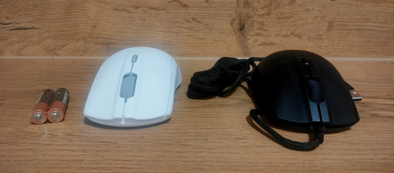 SteelSeries Rival 3 Gen 2 i Rival 3 Gen 2 Wireless Białą bezprzewodowa mysz komputerowa z przyciskami i rolką znajduje się pośrodku, obok niej po lewej stronie leżą dwie baterie AA, z prawej strony czarna przewodowa mysz komputerowa z widocznym kablem USB, całość na drewnianym tle.