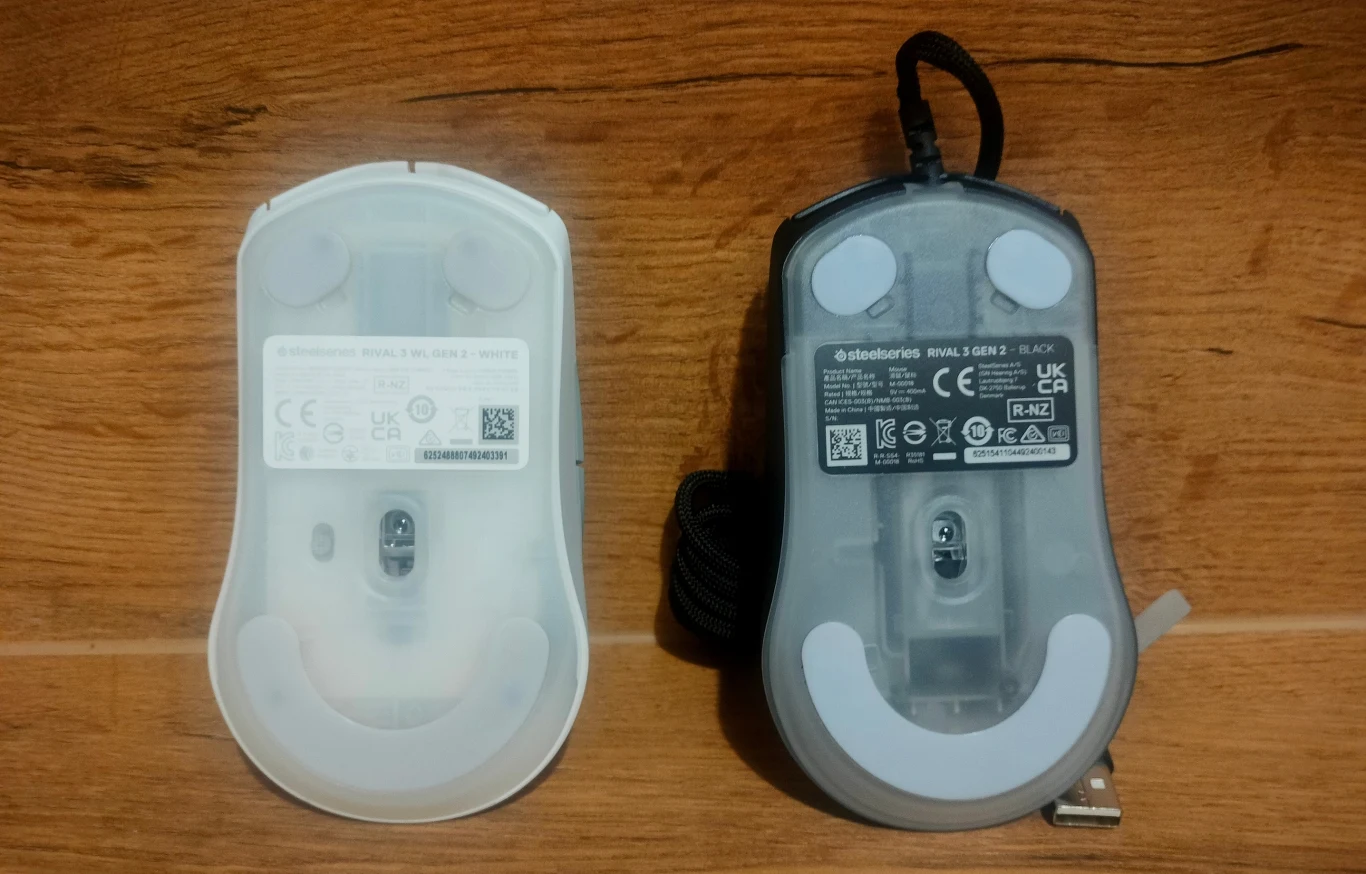 SteelSeries Rival 3 Gen 2 i Rival 3 Gen 2 Wireless Dwa przewodowe myszy komputerowe pokazane od spodu na drewnianej powierzchni, widoczne czarne i białe ślizgacze oraz etykiety z informacjami technicznymi i kodami QR.
