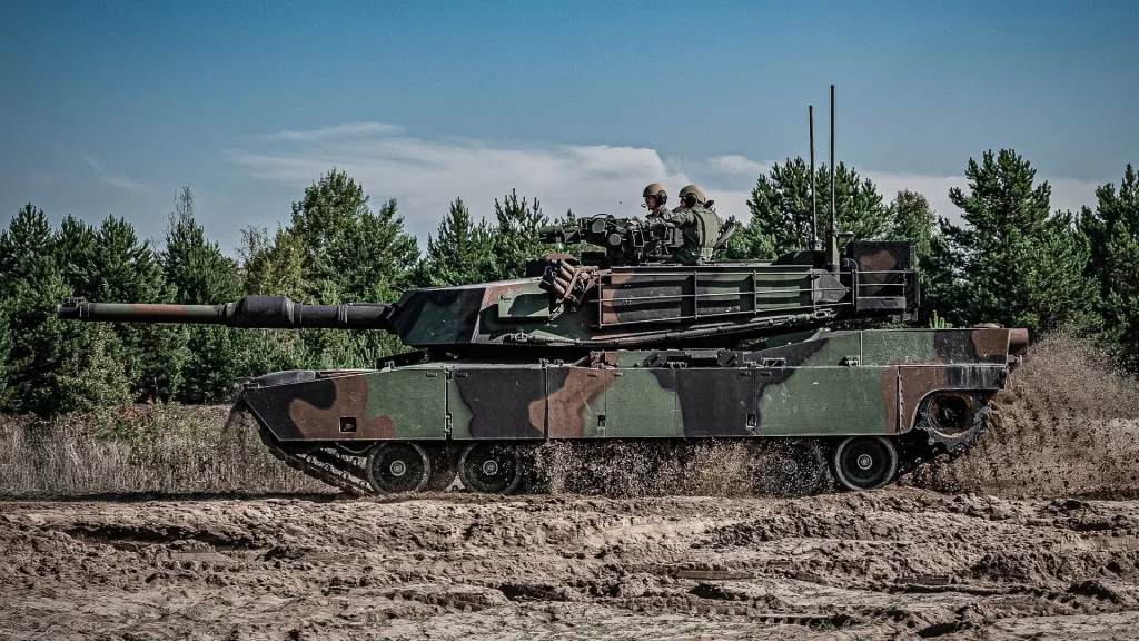 Czołg w kamuflażu porusza się po błotnistym terenie, otoczony gęstymi drzewami, z widocznymi żołnierzami w wieżyczce.