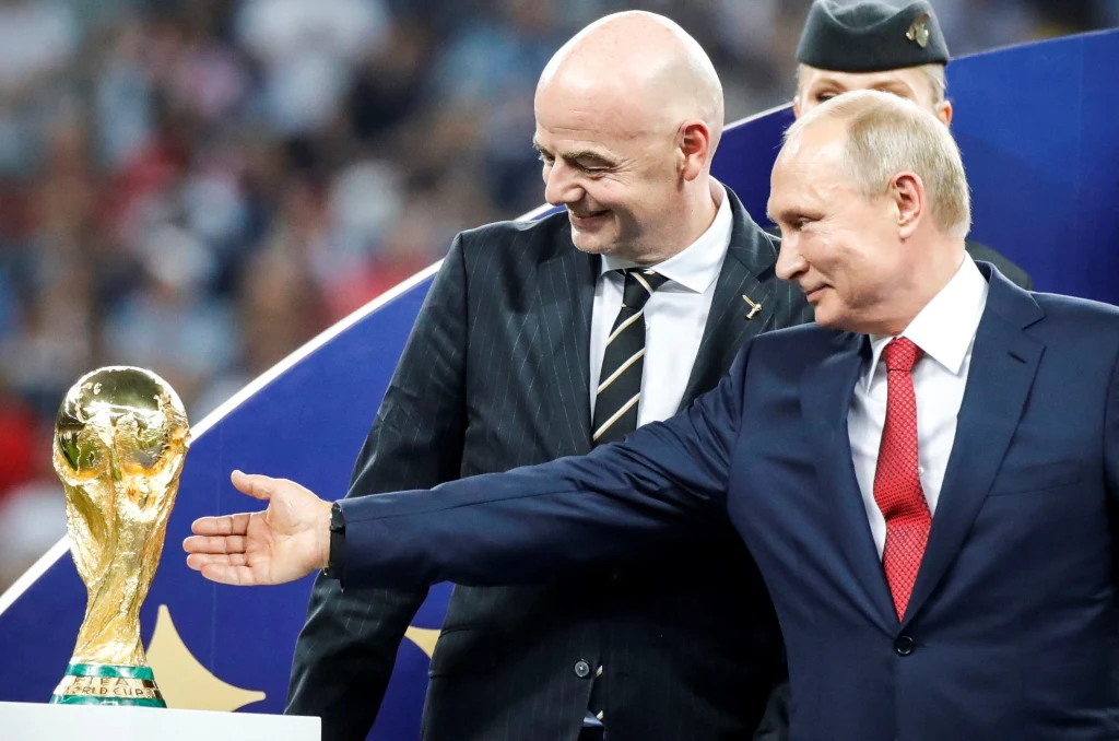 Szef FIFA Gianni Infantino i Władimir Putin, prezydent Federacji Rosyjskiej Dwóch mężczyzn w garniturach, jeden z nich wskazuje ręką na puchar świata w piłce nożnej. W tle rozmyte sylwetki widzów oraz człowiek w mundurze.