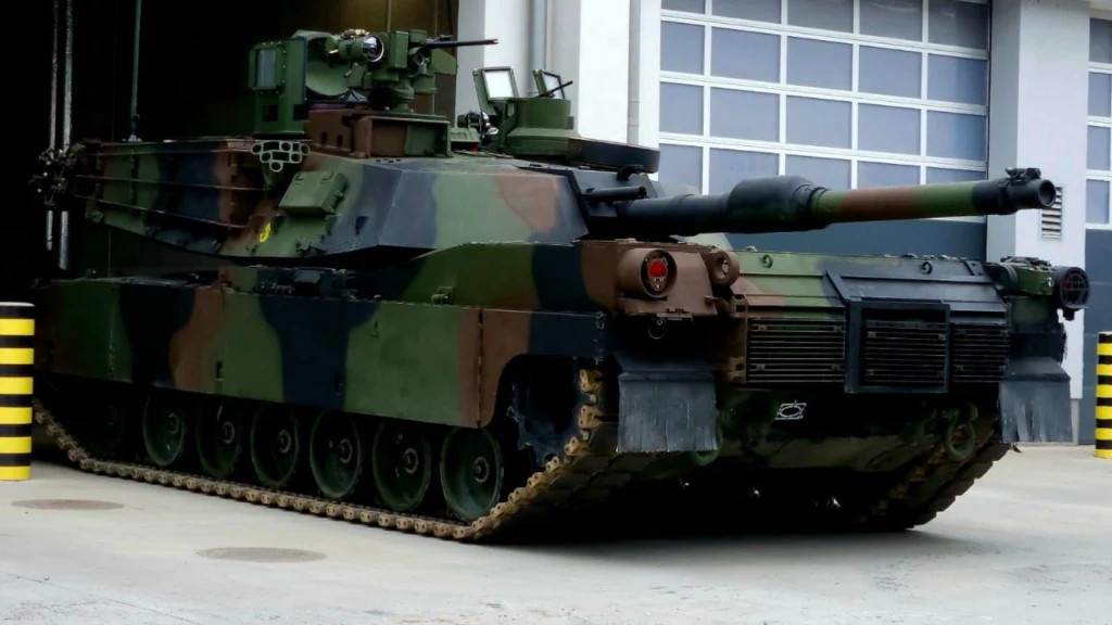 Czołg wojskowy w kamuflażu stoi zaparkowany przed dużym budynkiem z segmentową bramą. Widoczna armata skierowana jest lekko w bok, z tyłu czołgu znajdują się siatki ochronne, wyraźny gąsienicowy napęd oraz detale techniczne pancerza.