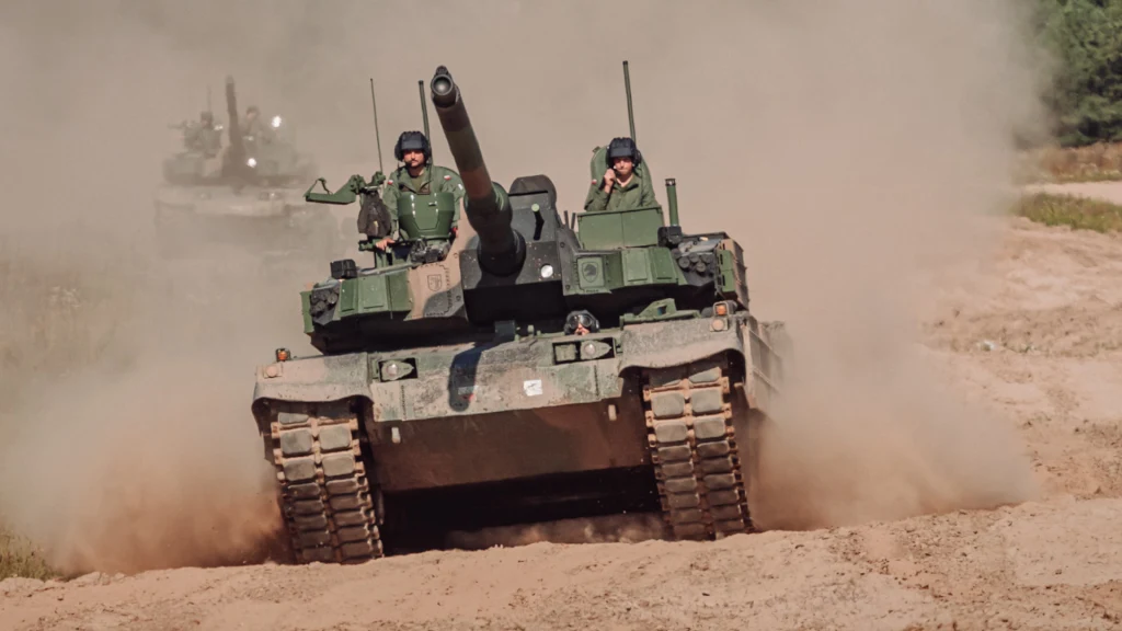 Czołg poruszający się po piaszczystym terenie z dwoma żołnierzami w wieżyczce, unoszący za sobą chmurę kurzu, w tle widoczny drugi czołg na poligonie.