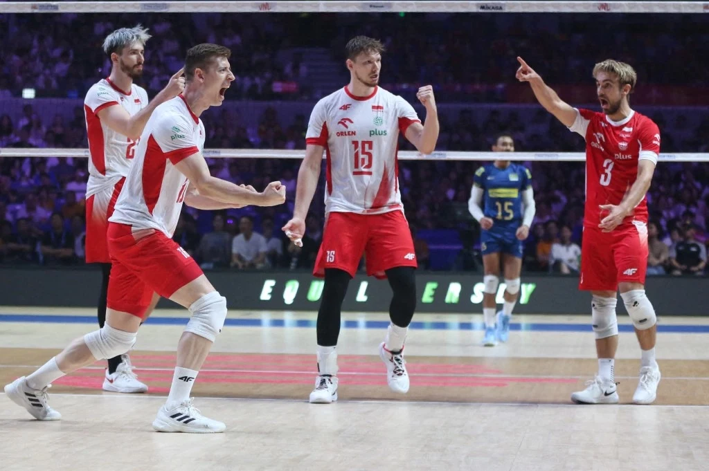 Grupa polskich siatkarzy w białych i czerwonych strojach świętuje zdobycie punktu na boisku, w tle widoczni zawodnicy drużyny przeciwnika oraz siatka.